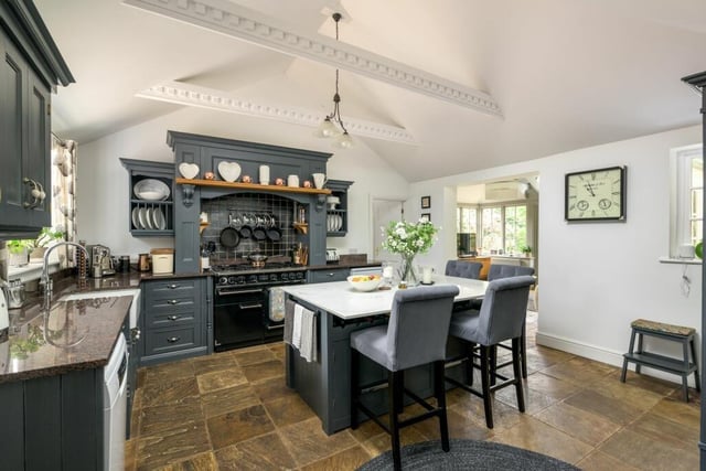 The kitchen has a modern farmhouse feel, with painted wooden units, mottled granite worktops, a central quartz-topped island, ceramic butler sink, stone tiled flooring, vaulted ceiling, and fireplace with a log burner.